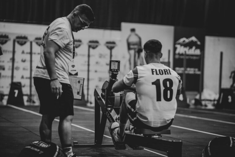 Atleta en una máquina de remo durante una competición, acompañado por un entrenador supervisando su rendimiento.