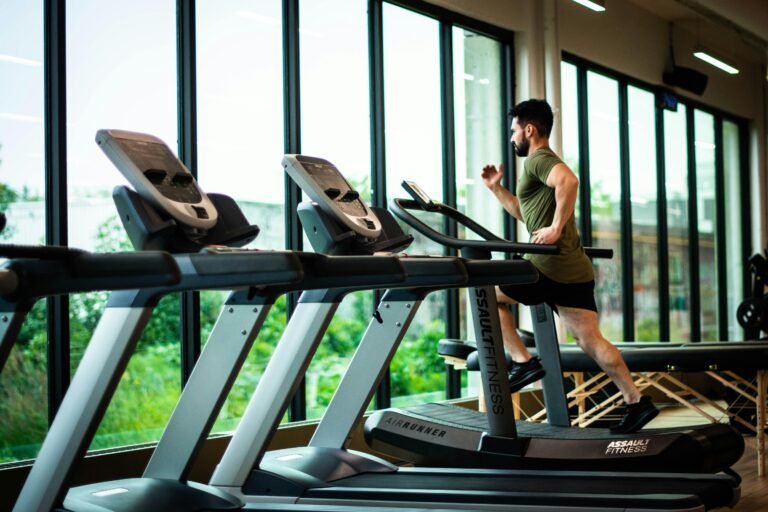 Un joven se ejercita en una cinta de correr en un gimnasio moderno con grandes ventanales y luz natural.